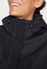 Schwarze Jacke mit hohem Kragen und verstellbaren Kordelzügen, glatter Textur und einer Frontleiste. Minimalistisches Design ohne sichtbare Muster.