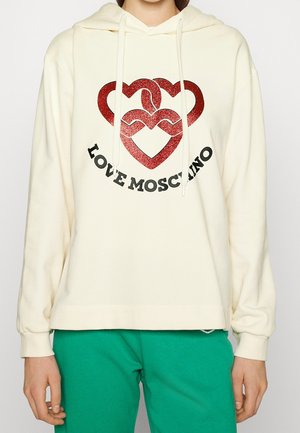Cremefarbener Hoodie mit roten, glitzernden, ineinander verschlungenen Herzen und schwarzem "LOVE MOSCHINO"-Schriftzug, kombiniert mit grüner Hose, Vorderansicht, die Hände locker an den Seiten.