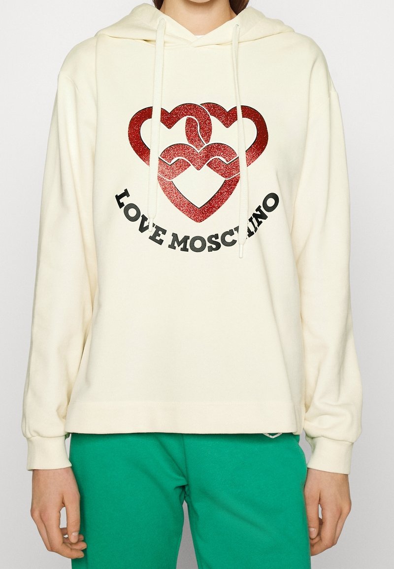 Sweat à capuche crème avec des cœurs entrelacés rouges pailletés et le texte noir « LOVE MOSCHINO », porté avec un pantalon vert, vue de face, les mains relaxées le long du corps.