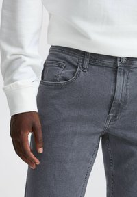 Vaqueros de mezclilla gris con un corte slim, diseño clásico de cinco bolsillos y cierre de botón. Combinados con una camisa blanca de manga larga.