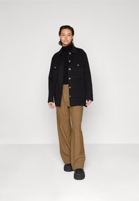 Schwarze oversized Jacke mit mehreren Fronttaschen, kombiniert mit einem schwarzen Rollkragenpullover und braunen weit geschnittenen Hosen. Schwarze Plateauschuhe runden den Look ab.