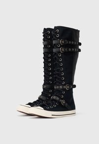 Hohe schwarze Schnürsneaker mit weißen Gummisohlen und mehreren schwarzen Schnallenriemen im Waden- und Knöchelbereich.
