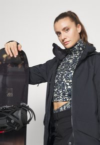 Veste noire sur un col roulé à motifs, avec un snowboard équipé de fixations. Textures et couleurs marquées dans les vêtements et l'équipement.