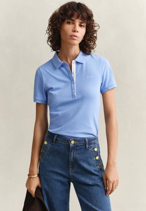 Femme aux cheveux bouclés portant un polo bleu clair et un jean taille haute bleu avec des boutons dorés, debout devant un fond clair.