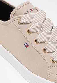 Beiger Leinwand-Sneaker mit weißer Sohle, gestreiften beige-weißen Schnürsenkeln, goldfarbenen Ösen und kleinem Tommy Hilfiger Flaggenlogo an der Seite.