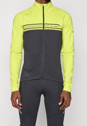 Mann trägt eine schwarze und neongelbe langärmlige Fahrradjacke und schwarze Fahrradhosen, steht vor einem einfarbigen Hintergrund.