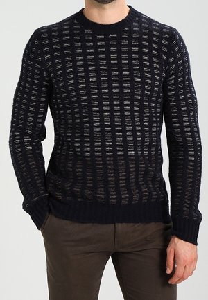 Mężczyzna ubrany w czarny dziany sweter z pionowymi paskami o otwartym splocie oraz brązowe spodnie, stojący z jedną ręką w kieszeni.