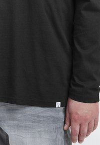 Black long-sleeve shirt made of soft fabric, with a side logo tag visible. Hand rests on grey distressed jeans. Texture appears smooth.