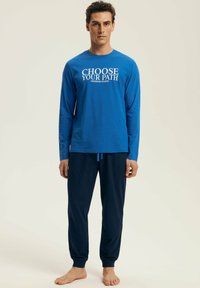 Man standing barefoot wearing a blue long-sleeve shirt with "CHOOSE YOUR PATH" text and dark navy jogger pants against a plain background.