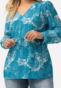 Blusa turchese a maniche lunghe con scollatura a V, vita regolabile con cordino e motivo floreale bianco su uno sfondo testurizzato.