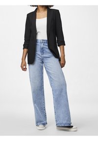 Femme portant un jean large bleu clair, un débardeur blanc, un blazer noir avec manches retroussées, et des baskets noires à semelles blanches.