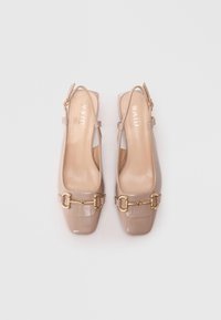 Beige patent leather slingback-kengät, joissa on neliön muotoiset varpaat ja kultaiset hevosketju-yksityiskohdat, aseteltuna vierekkäin vaalealla taustalla.