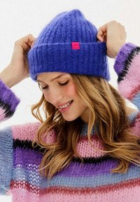 Fialová žebrovaná pletená čepice s ohrnutým lemem a malou růžovou značkovou etikou. Nnošená s barevným pruhovaným svetrem. Měkká textura a těsný střih.
