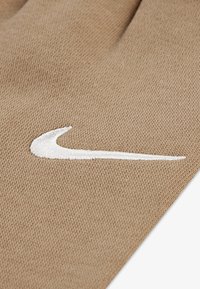 Beigen värinen neulottu käsine, jossa on näkyvä valkoinen kirjailtu Nike Swoosh -logo. Kangas on teksturoitu tiheällä kudontakuviolla.