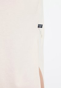 T-shirt beige chiaro con una fessura laterale e un'etichetta nera. Realizzato in tessuto morbido con una superficie liscia, presenta una silhouette rilassata.