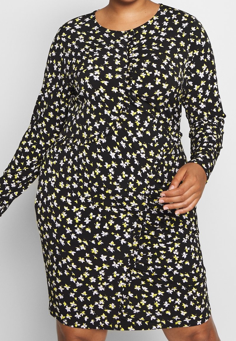 Femme portant une robe noire à manches longues avec un petit imprimé floral blanc et jaune et un col rond.