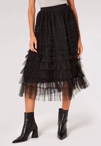 Jupe en tulle noir avec des volants superposés, longueur mi-mollet, taille élastique, associée à des bottines noires brillantes à bouts pointus.