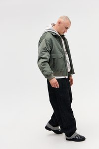 Casaco bomber verde-oliva sobre um hoodie branco, combinado com jeans largos pretos e sapatos pretos com detalhes cinzas. Design simples, cores neutras.