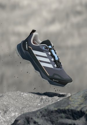 Turistická obuv GORE-TEX s tmavomodrým sieťovaným zvrškom, bielym pruhovým detailom, čiernymi akcentmi a robustnou sivou podrážkou, zobrazená vo vzduchu nad skalou.