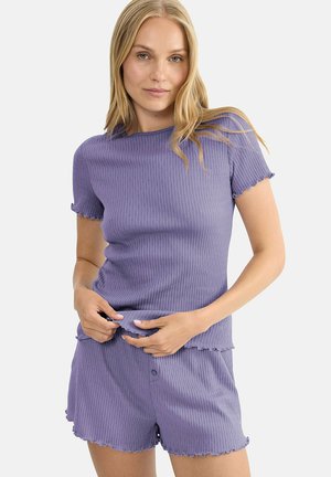 Femme portant un haut court à manches courtes côtelé lavande et un short assorti avec des bords ondulés, ajustant sa chemise sur un fond uni.