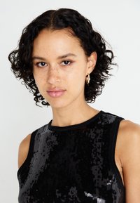 Top de tirantes negro con lentejuelas, diseño sin mangas, escote redondo y textura suave. La modelo tiene el cabello rizado y lleva pendientes de aro.
