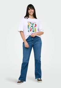 Camiseta de algodón blanca con un diseño gráfico en verde y rojo y la palabra "PIMENTÓN". Lleva puesta con unos vaqueros de campana azules y zapatillas marrones.