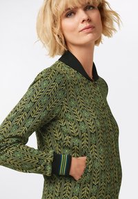 Vihreä kuvioitu takki mustalla taustalla, jossa on ribatut kaulus ja hihansuut sekä kaksi sivutaskua. Yksityiskohtainen kukkakuvio.