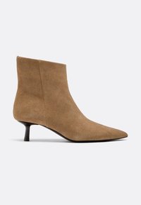 Brun ruskindsankelstøvle med spids tå og lav, slank hæl mod ensfarvet lys baggrund.