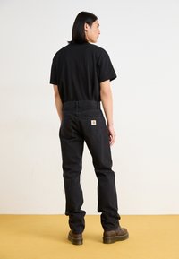 Svart kortermet t-skjorte kombinert med svarte denimjeans, med en logo på baksiden. Modellen har på seg brune støvler og står på en gul overflate.