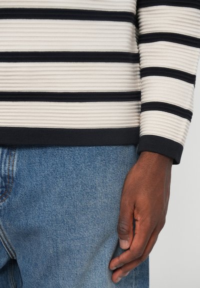 Puli s crew-izrezom s horizontalnim prugama u krem i mornarsko plavoj boji, s rebrastom teksturom. Uparen s plavim trapericama.