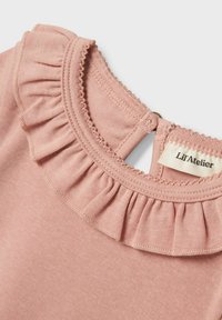 Gaišas rozā kokvilnas apģērbs, kuram ir volāna apkakle, aizdare ar pogu aizmugurē un birka ar uzrakstu "Lil'Atelier".