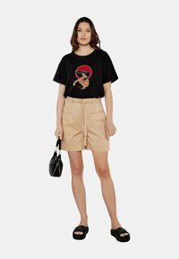 T-shirt grafico nero con un design circolare rosso, abbinato a pantaloni corti beiges a vita alta. Il modello porta una borsa nera e indossa sandali neri.