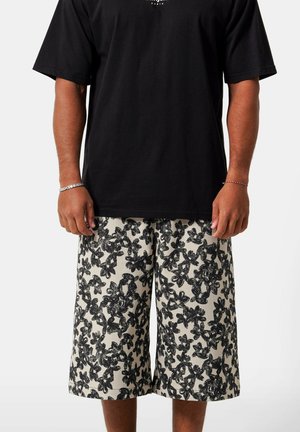 Persona con camiseta negra de manga corta y pantalones cortos sueltos hasta la rodilla con un patrón floral negro sobre fondo blanco.