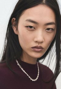 Prim-plan al unei persoane cu păr lung și negru, purtând un pulover bordo cu guler înalt și un lanț argintiu la gât. Fundal neutru.