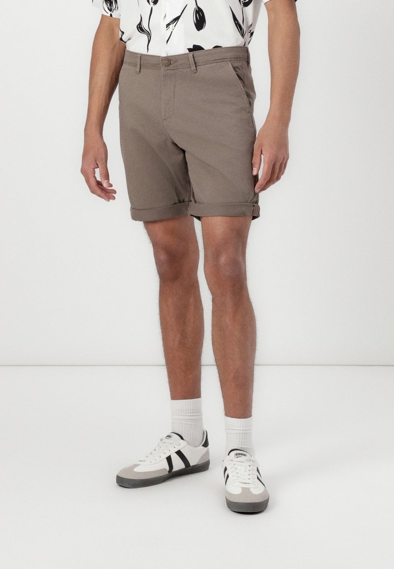 Homme portant un short marron remonté, des chaussettes blanches, des baskets blanches et noires, et une chemise blanche avec des motifs de feuilles noires.