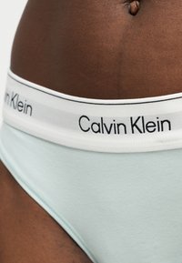 Gros plan sur le bas du torse d'une personne portant un sous-vêtement Calvin Klein bleu clair avec une ceinture blanche et un logo noir.