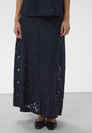 Femme portant une jupe longue en dentelle bleu marine avec un motif floral et des chaussures plates noires à bout pointu.