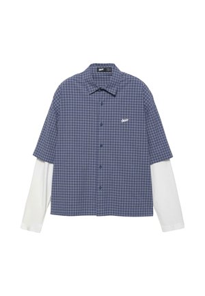 Camicia a maniche corte a quadri blu e bianchi indossata sopra una camicia bianca a maniche lunghe, con bottoni sul davanti e piccolo logo bianco sul petto.