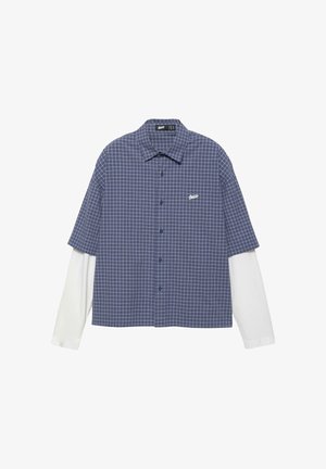 Camicia a maniche corte a quadri blu e bianchi indossata sopra una camicia bianca a maniche lunghe, con bottoni sul davanti e piccolo logo bianco sul petto.