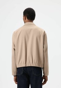 Beige jas met ritssluiting, gemaakt van een gladde, gestructureerde stof, met elastische zoom enstaande kraag. Draagt over een donkerblauwe spijkerbroek, gezien van de achterkant.