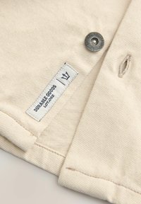Beige denim stof met een label waarop "DURABLE GOODS" staat en een genaaide knop. Beschikt over een schone, eenvoudige ontwerp en nauwkeurige stiksels.
