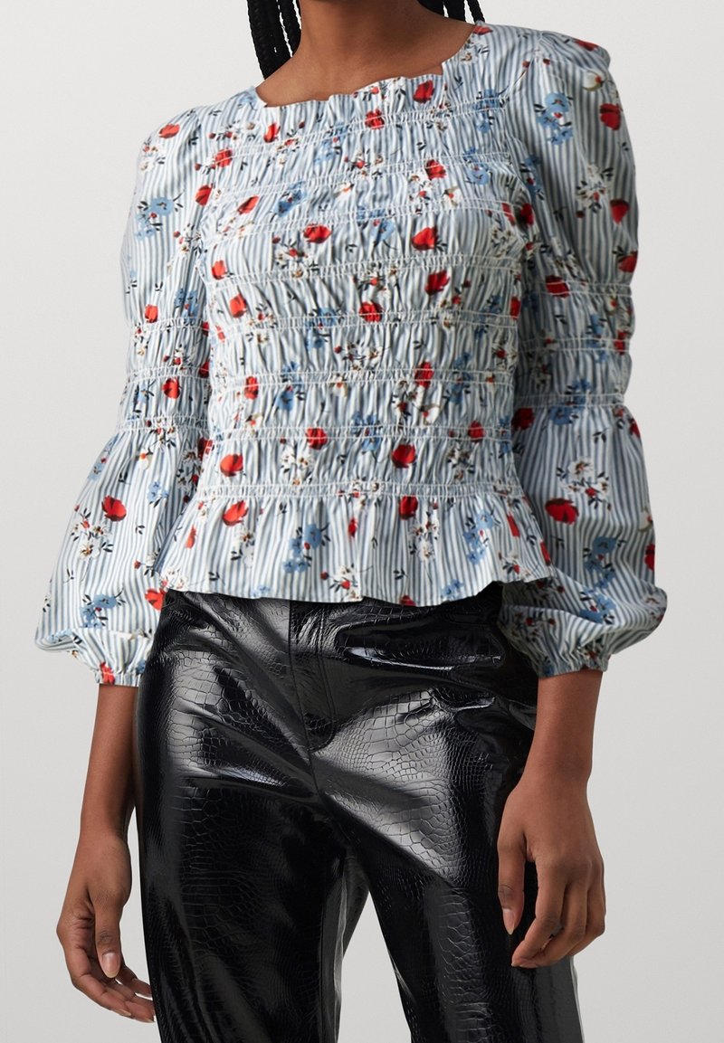 Femme portant un chemisier fleuri rayé bleu et blanc avec des manches bouffantes et un pantalon noir brillant texturé.