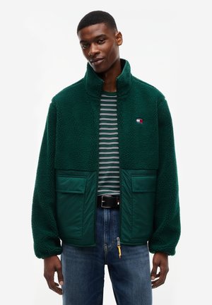 Man draagt een groene sherpa fleecejas met twee grote voorzakken, gestreept overhemd en blauwe spijkerbroek tegen een witte achtergrond.