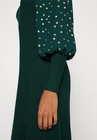 Groene jurk met aansluitend lijfje, opgeblazen mouwen van donkergroene stof met gouden stippen. Geribbelde manchetten en een gladde textuur.