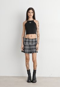 Black cropped halter top with a logo, paired with a black and white plaid pleated skirt. Model wears black chunky ankle boots.