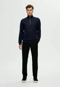 Marineblauer Zip-Neck-Pullover mit langen Ärmeln, strukturiertem Strick, kombiniert mit schwarzen Hosen und schwarzen Sneakers, vor einem einfarbigen Hintergrund.