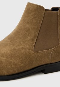 Botines de ante marrón con paneles laterales elásticos y un tacón bajo, con una textura suave y un diseño moderno y elegante.