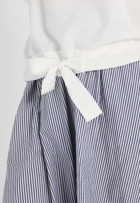 Biała bluzka związaną kokardą w talii, na granatowo-białą spódnicę w paski z plisami, podkreślającą fakturę i wzór materiału.