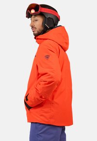 Feloranjeskijack met capuchon, voorzien van zwarte accenten en logo op de mouw. Glad materiaal, ontworpen voor gebruik in koude weersomstandigheden.