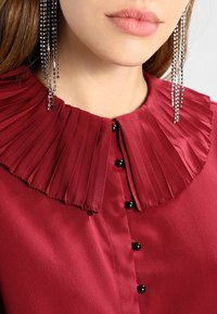 Blusa cremisi con colletto a pieghe e rialzato, caratterizzata da bottoni neri lungo la parte anteriore e un tessuto strutturato. Orecchini scintillanti completano il look.